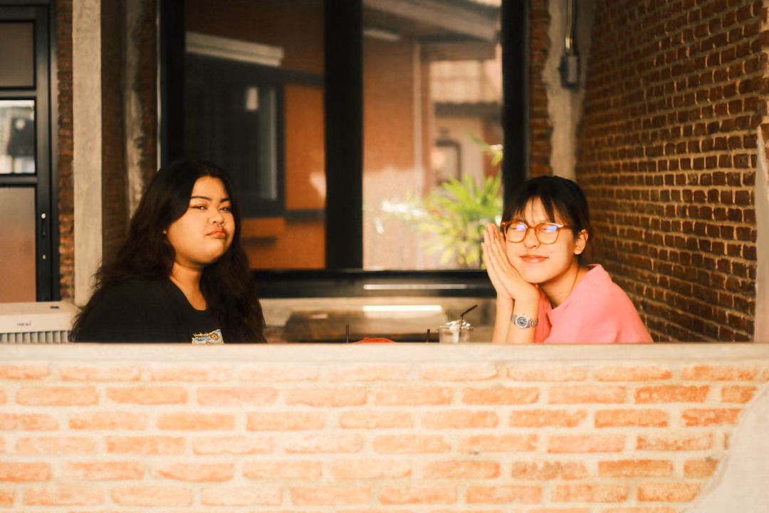 Two women at a window table behind a brick wall, one resting her face on her hands, the other looking sideways