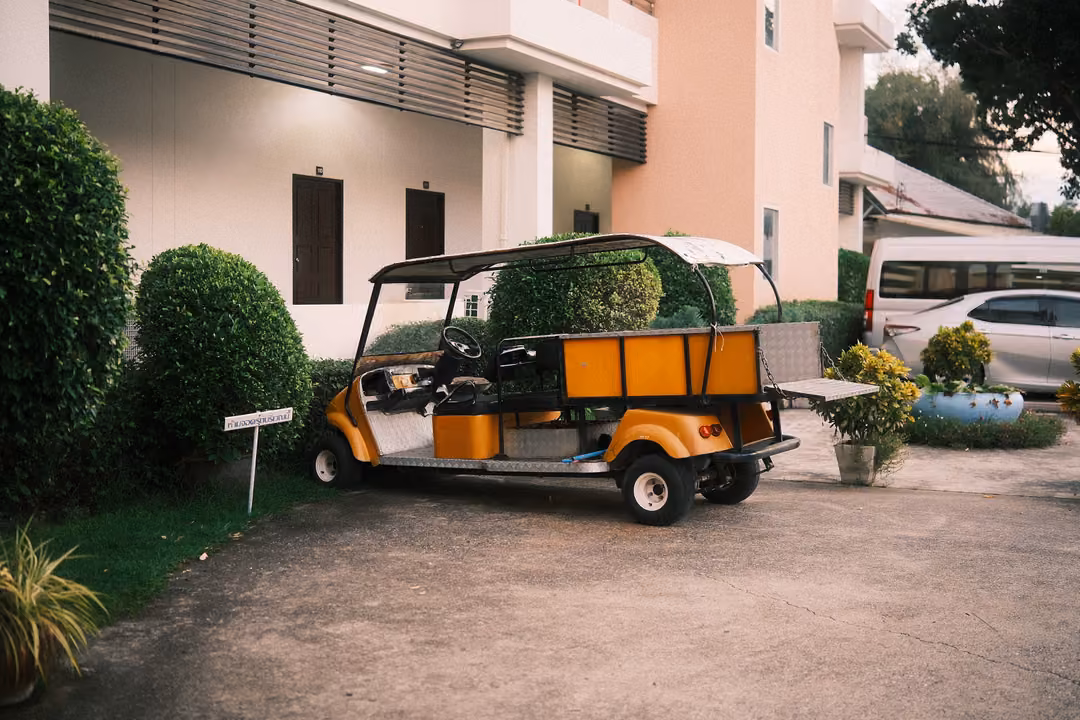 Orange and black golf cart parked on a driveway beside trimmed hedges and a pale pink building