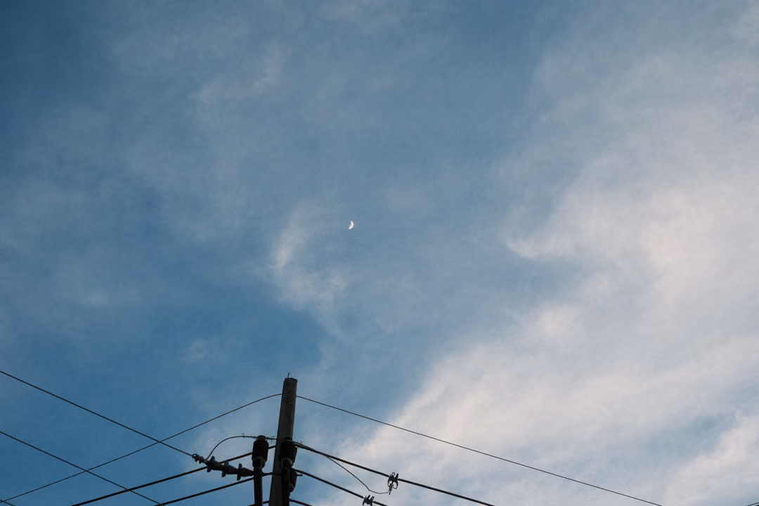 Thin crescent moon in a blue cloudy evening sky above a power pole and wires