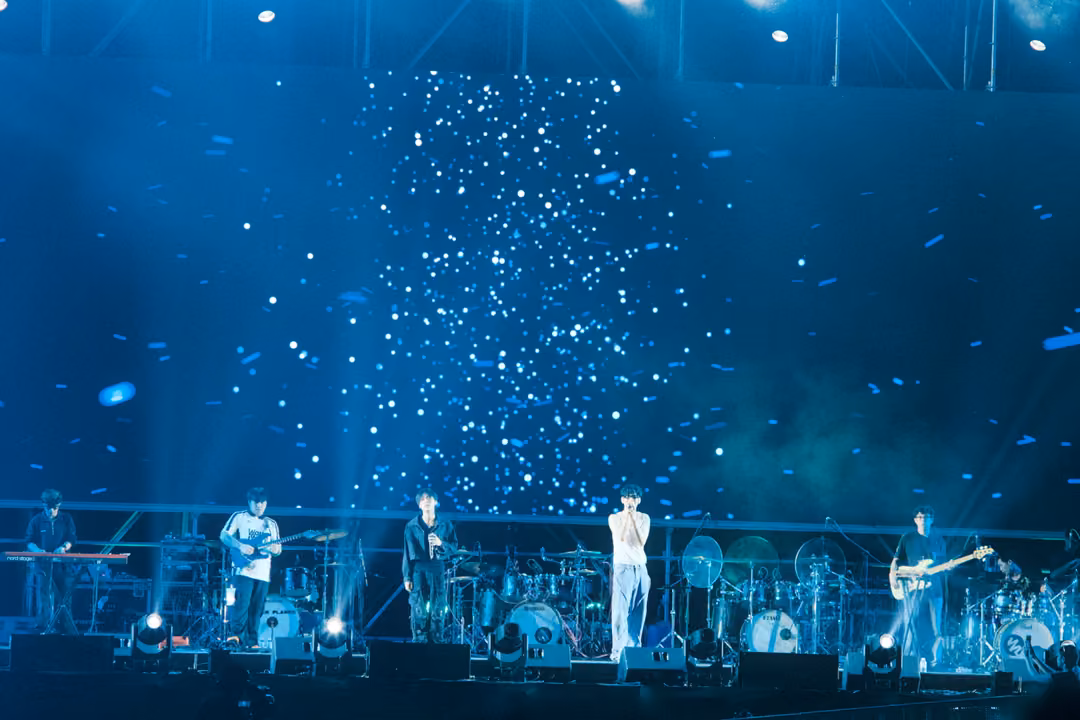 Band performing on stage beneath a screen of falling white flecks, blue haze and lights around them.