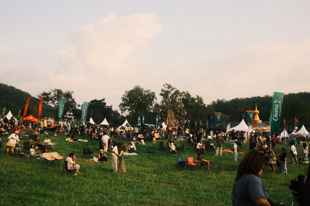 A large crowd scattered across a grassy field with white tents, flags and trees under a cloudy sky.