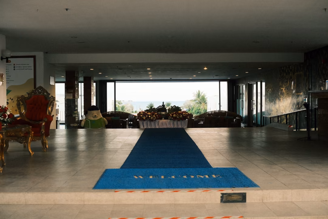 Spacious hotel lobby with a blue carpet runner and WELCOME mat leading toward glass doors and a flower display