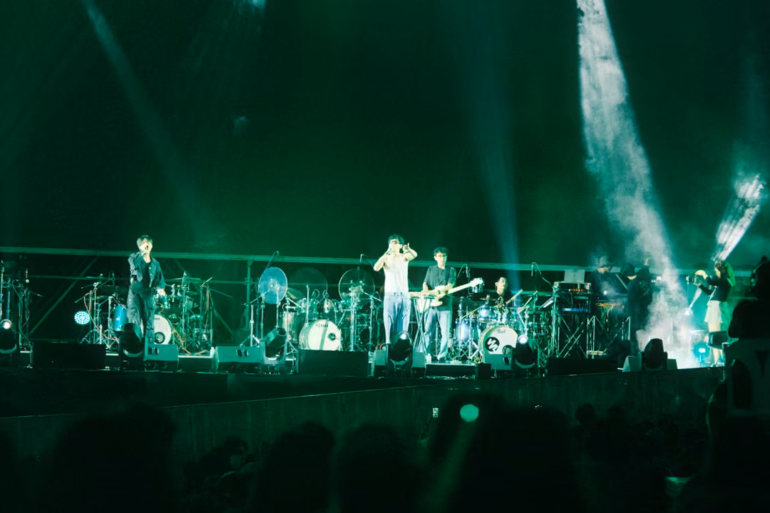 Band on a stage bathed in deep green light, white smoke jets rising at right, crowd silhouetted below.