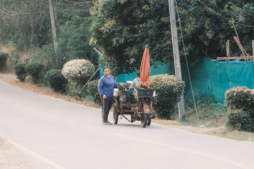 Person in a blue top pushing a loaded cart with a closed red umbrella along a quiet roadside