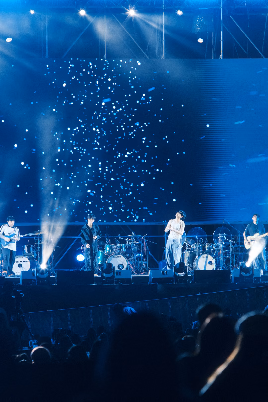 Vertical view of the stage with white light beams rising into a screen of white flecks, crowd silhouettes below.