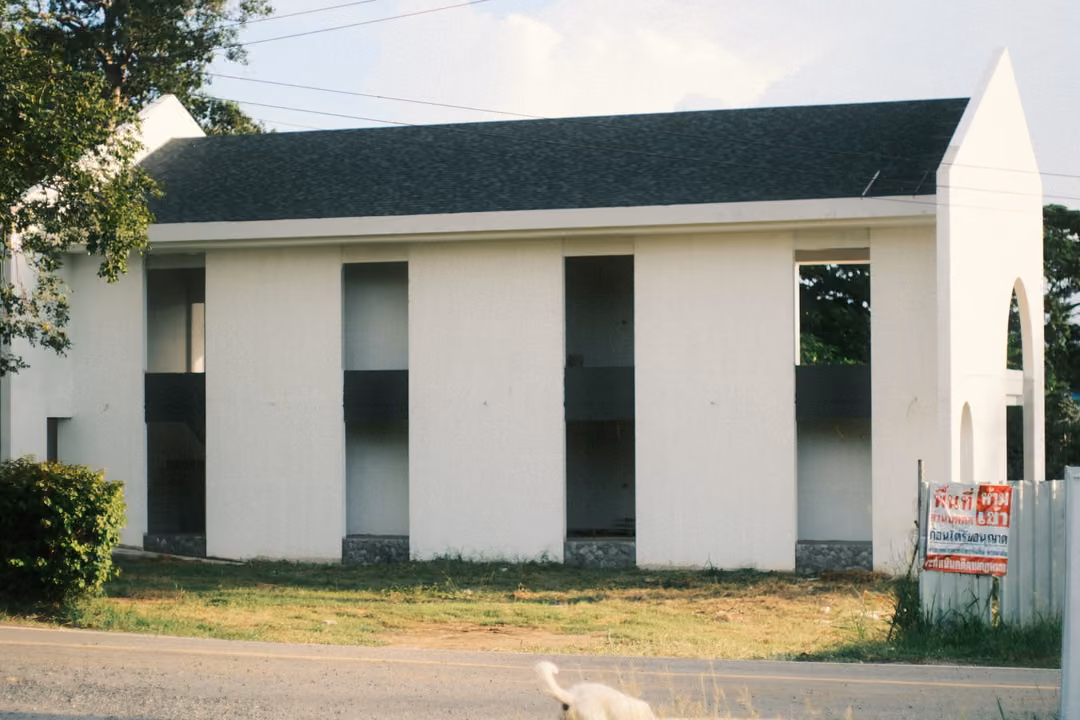 White two-storey building with tall openings and a gabled end, a red sign by the fence, a white bird by the road