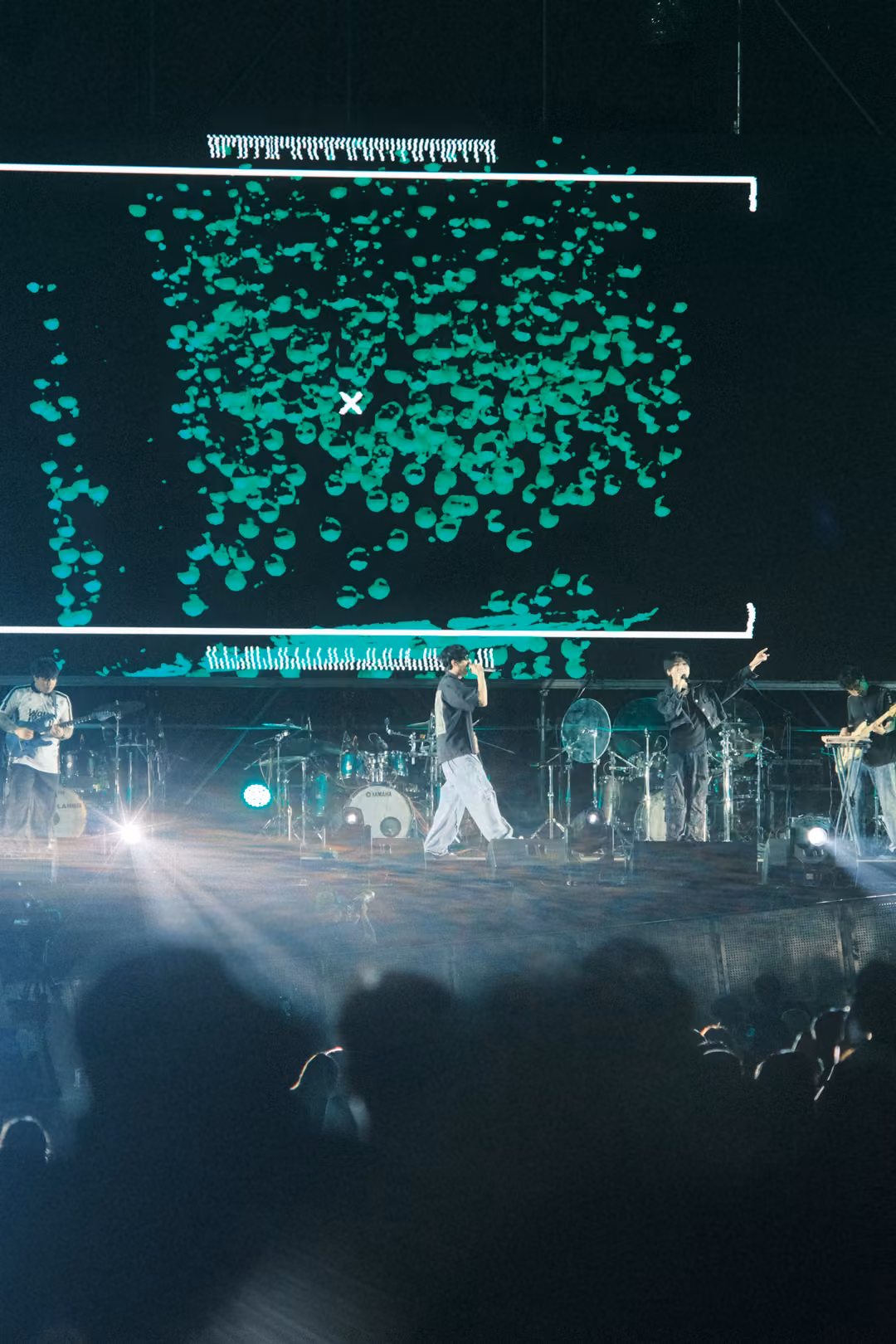 Tall view of a stage under a screen full of green shapes inside a white bracket, white light flaring across the crowd.