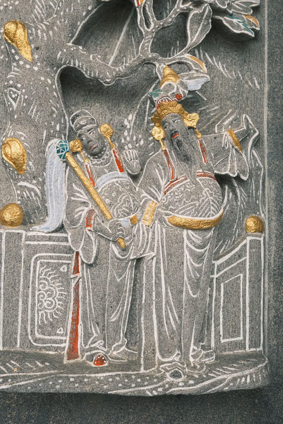Grey stone relief of two robed figures under a flowering tree, accented with gold, red and white paint