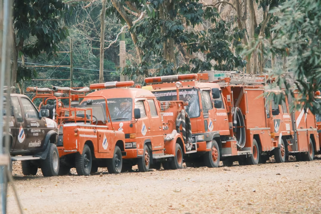 Row of orange-red fire and rescue trucks parked along a leafy dirt road under trees