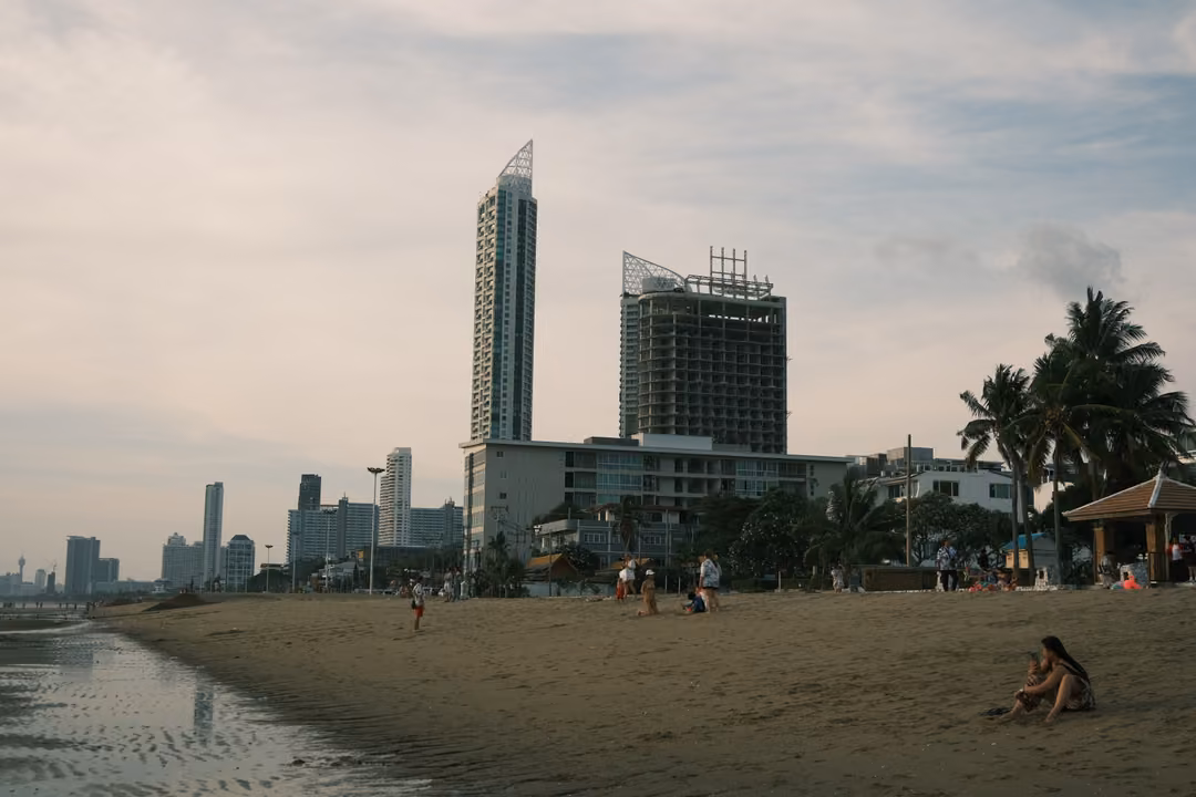 A beach with scattered people below tall city towers and palm trees, under a pale cloudy sky