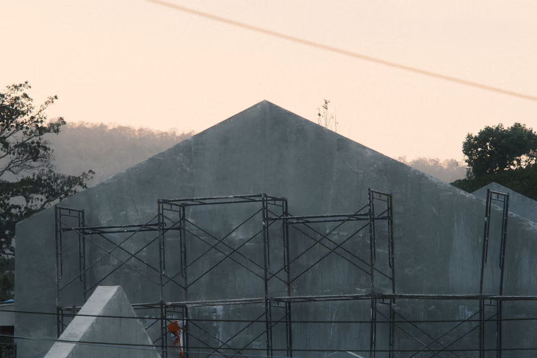 Grey concrete gabled building wrapped in metal scaffolding against a peach sky and distant hills