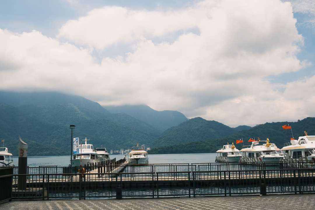 Railed lakeside pier with white tour boats and red flags, forested mountains beneath big white clouds