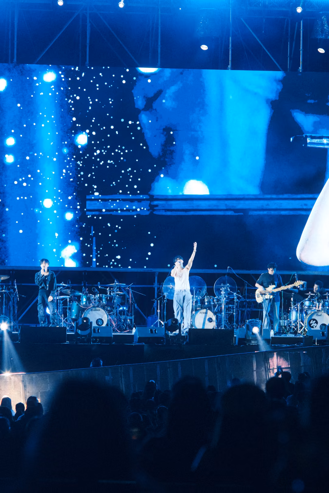 Vertical stage view with the singer raising one arm under a screen of white specks, guitarist and drums nearby.