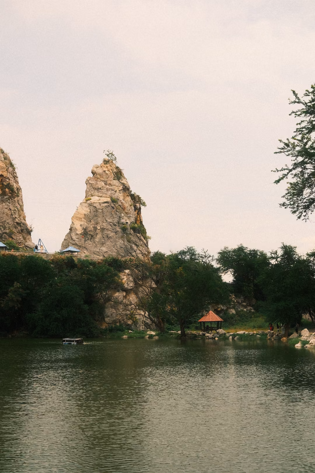 Pointed rock spire rising above trees and a still lake, a small pavilion on the shore and an overcast sky