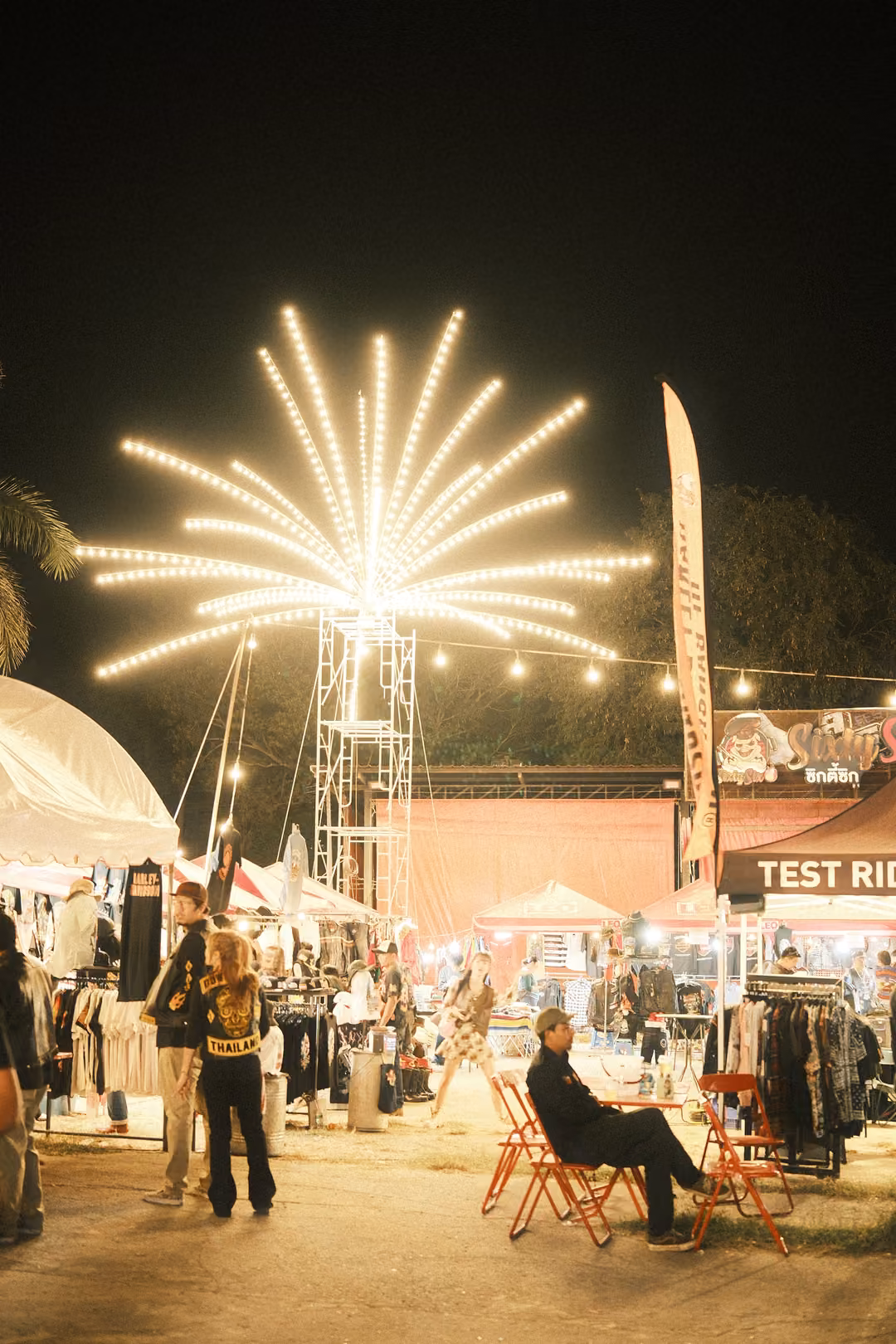 Starburst light tower above festival stalls with clothing racks, folding chairs and a Test Ride sign