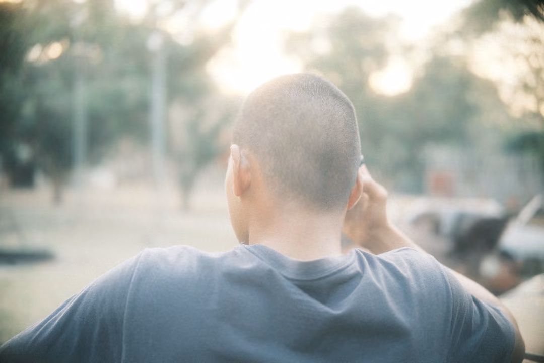 Back of a man's shaved head and shoulders in a grey t-shirt, hand at his ear, hazy trees and light