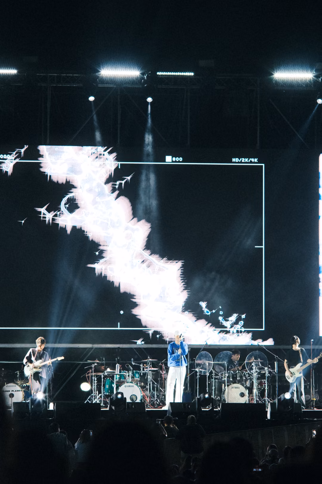 Vertical view of a screen with a white jagged shape in a framed box, the singer and guitarists performing below.