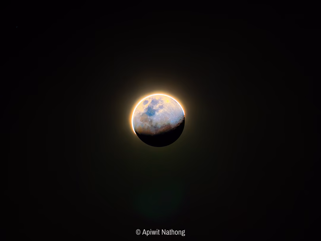 A moon partly lit, its shadowed part glowing faintly, with a bright golden rim against black space