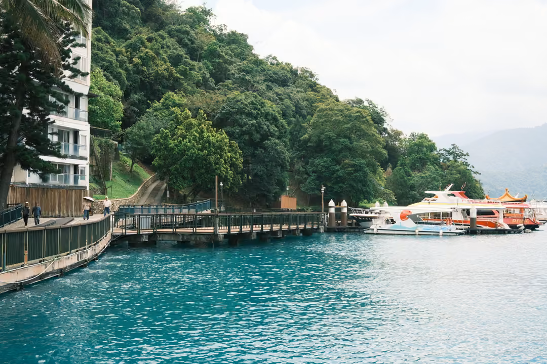 Turquoise lake water beside a wooded hillside, a boardwalk leading to boats moored at a small pier