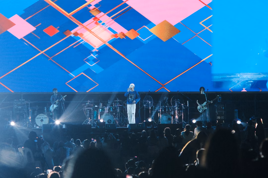 Band performing beneath bright blue and pink geometric graphics on screen, white beams lighting the stage edge.