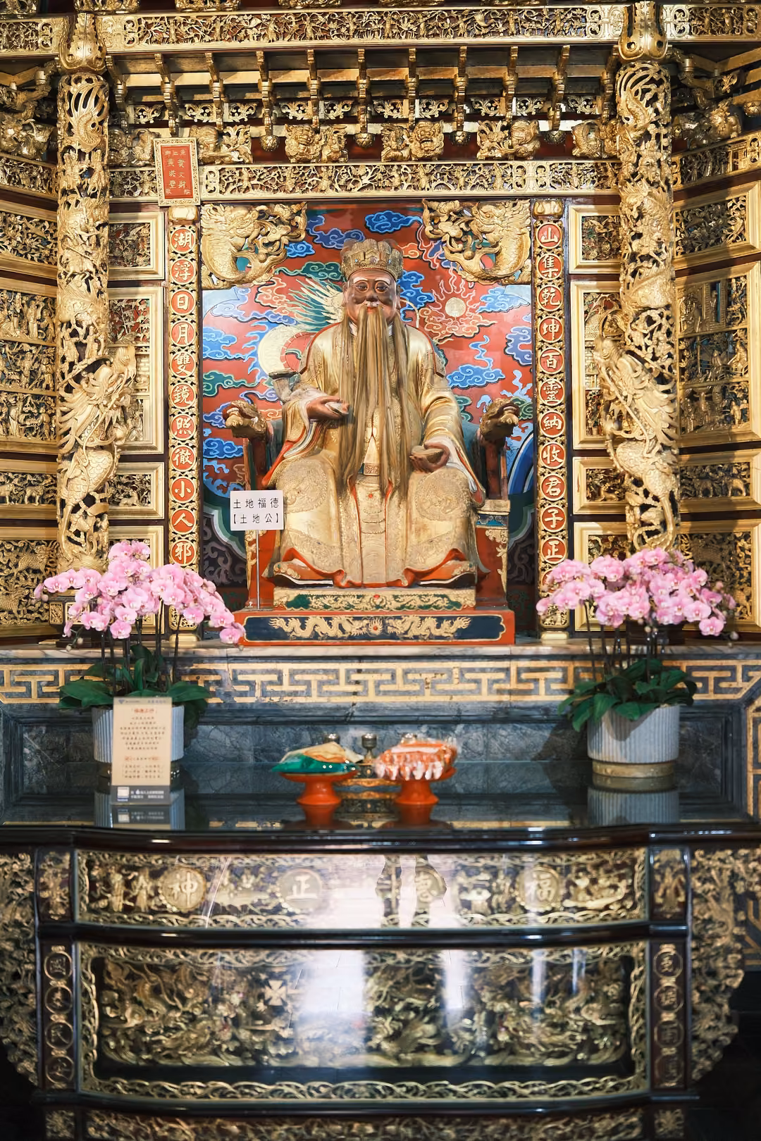 Gilded shrine with a seated figure with a long white beard in gold robes, pink orchids and offerings in front