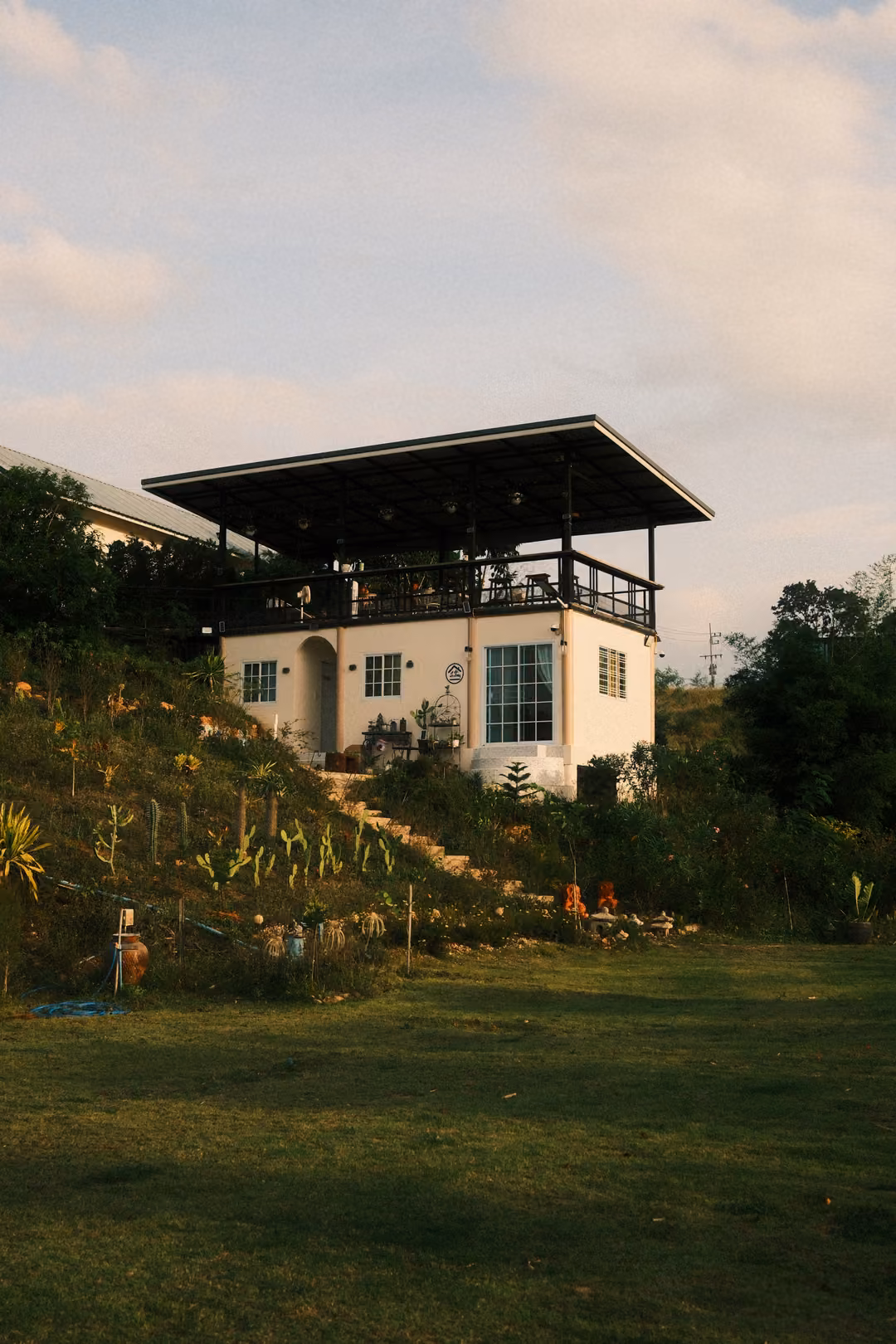 A white two-storey house with a roofed terrace on top, sitting on a grassy slope with steps and plants.