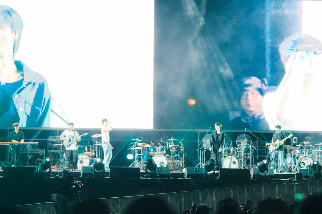 Band on stage below a screen that is mostly bright white with a figure at the left and a singer at the right.