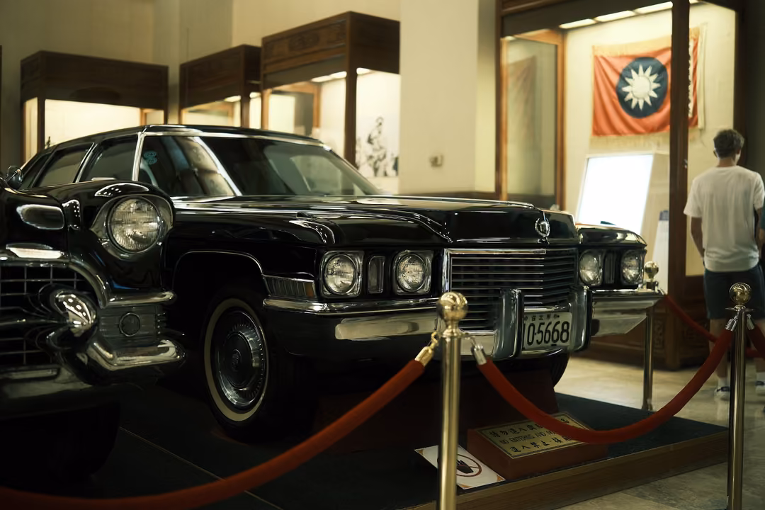 Black vintage limousine with quad headlights behind red ropes, a visitor in white facing a flag display