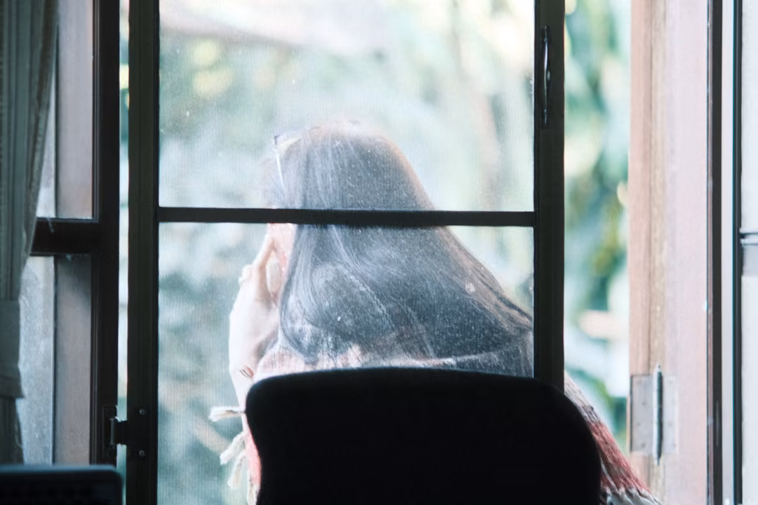 Person with long dark hair seen from behind at an open window, a chair in the foreground