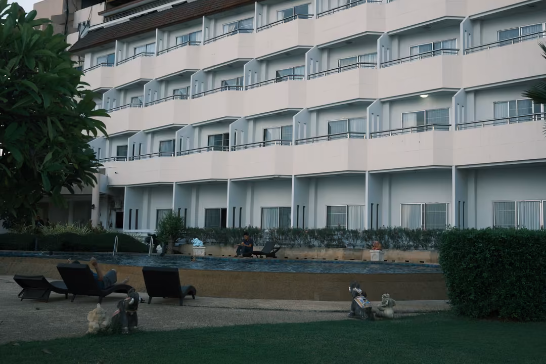 White multi-storey hotel with rows of balconies, a pool area with loungers and a lawn in front