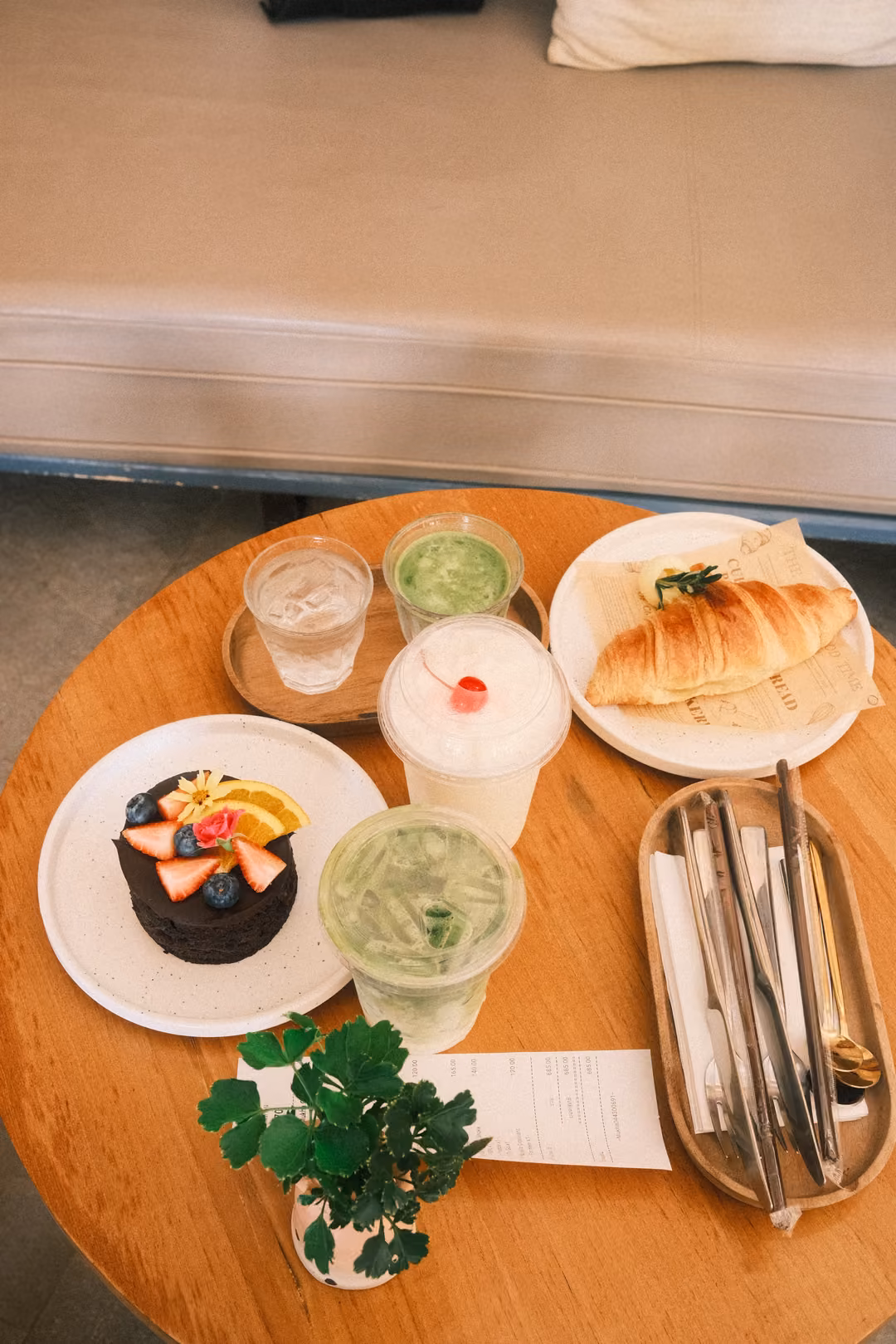 Round wooden table with a croissant, a small fruit-topped cake, matcha and milk drinks, and a small plant