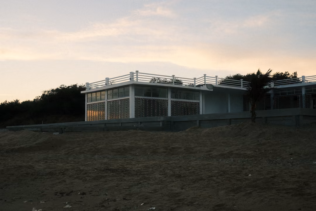 A white modern beachfront building with a railing on its roof, seen across dark sand under a pale dusky sky