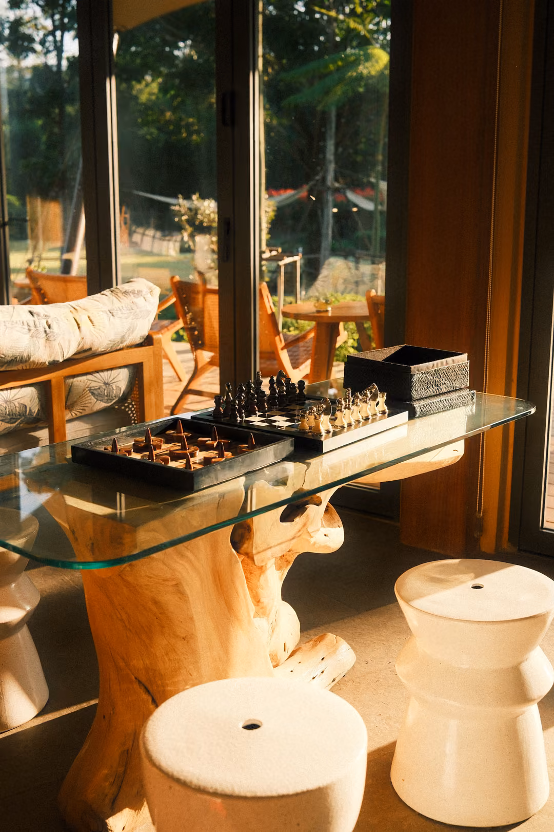 Vertical view of chess sets on a glass table with a carved wooden base, white stools and sunlit windows