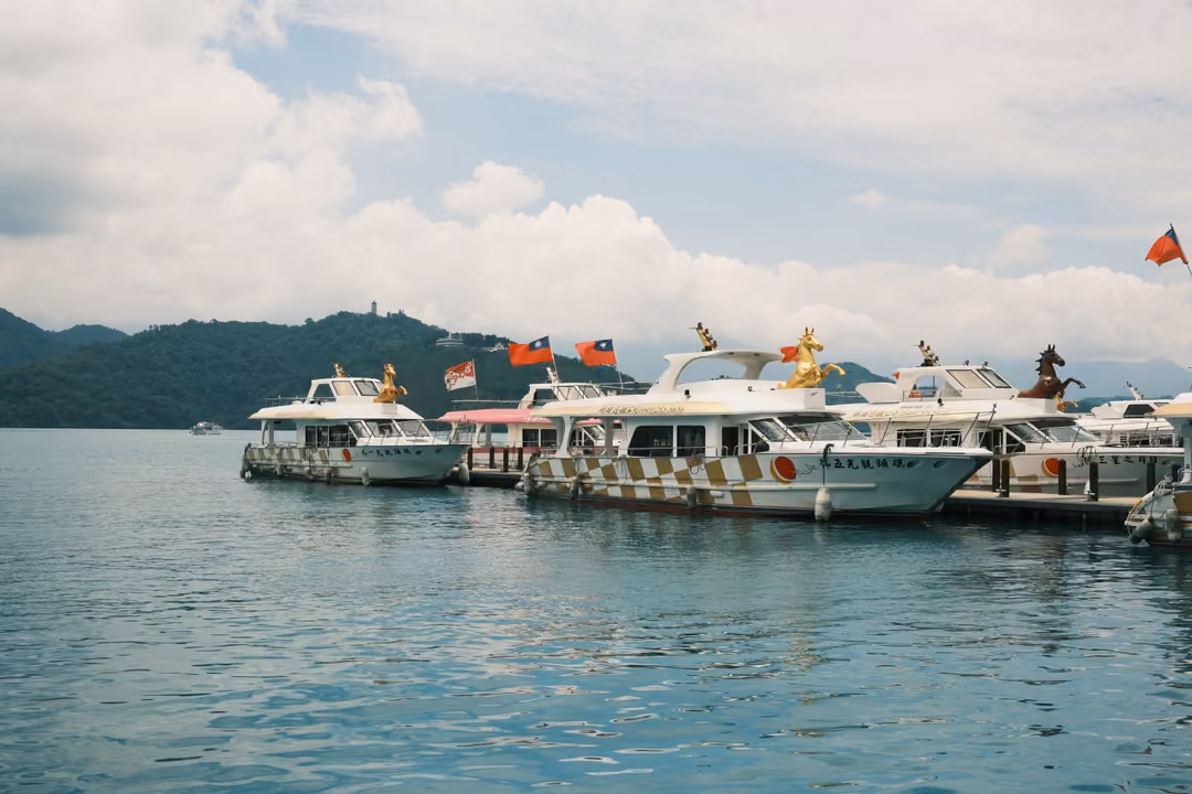 Tour boats with gold figurines and red flags moored at a pier on a calm lake, wooded hills behind