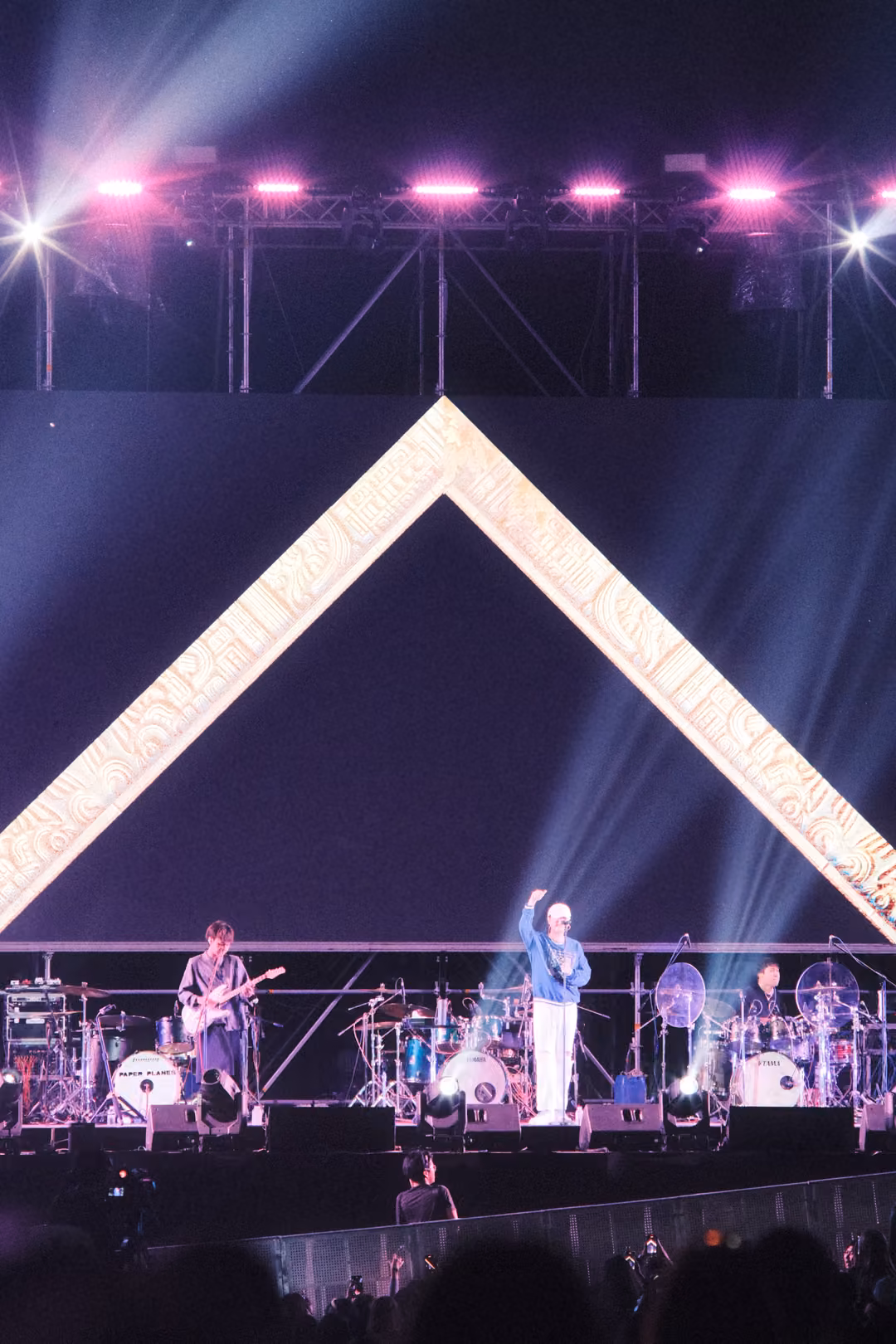 Vertical view of a singer with one arm raised under an empty gold-framed triangle, pink lights on the truss above.
