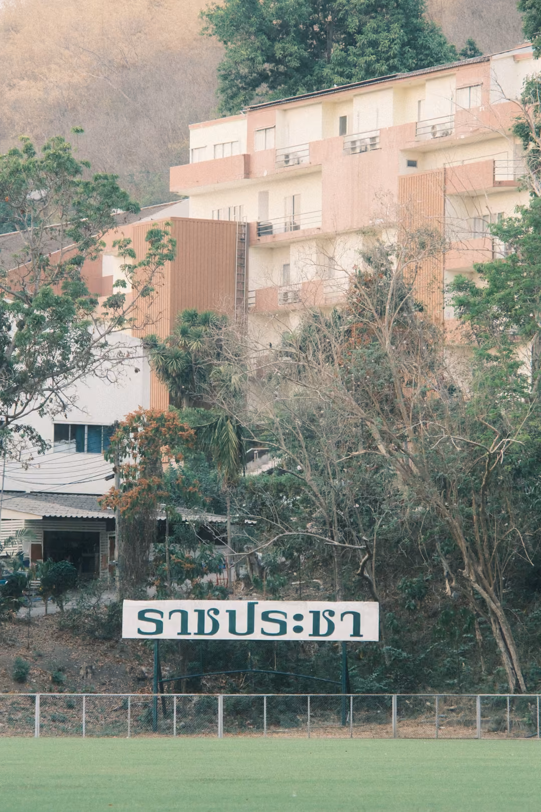 Peach and white multi-storey building on a hillside behind trees, a white sign with Thai lettering by a field