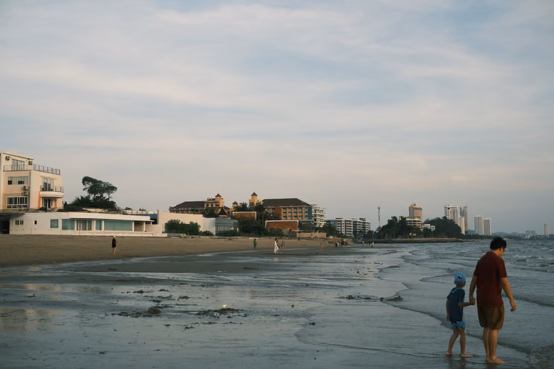 An adult and a small kid in a blue cap walking hand in hand along the waterline, buildings behind