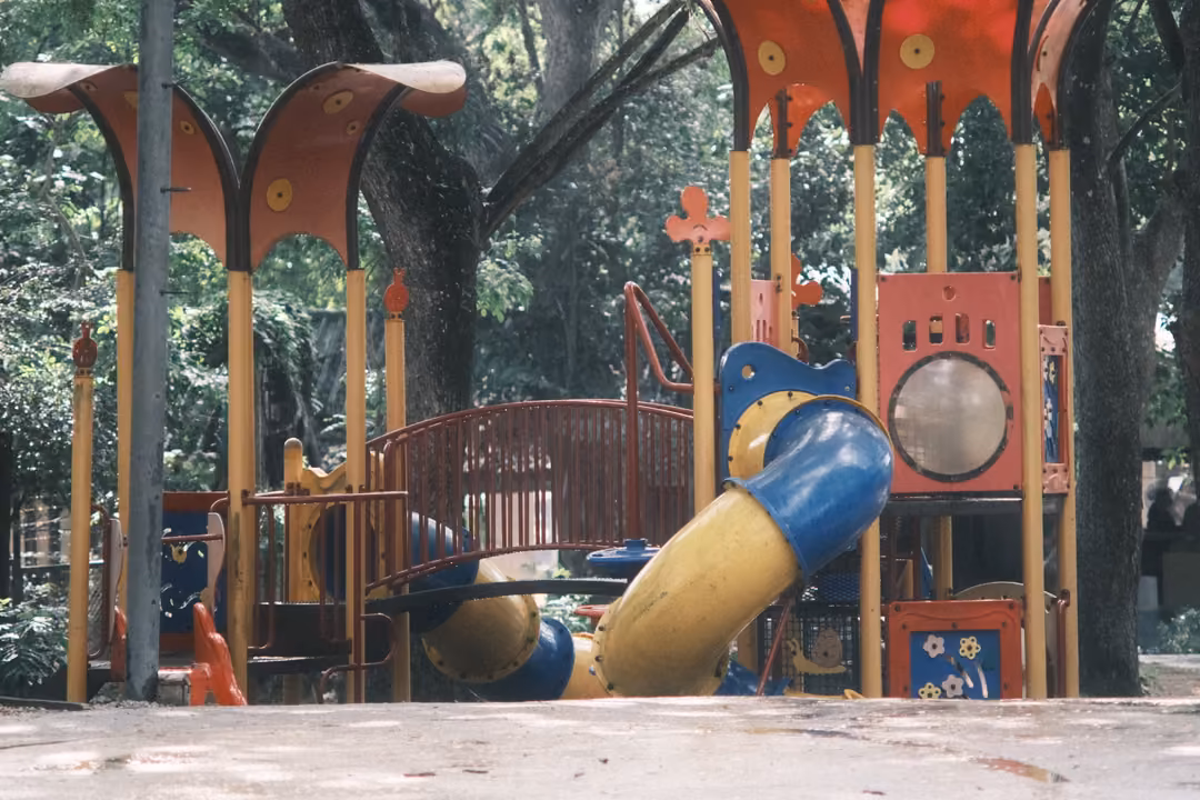 Empty outdoor playground with a yellow tube slide, red canopies and wooden posts under shady trees