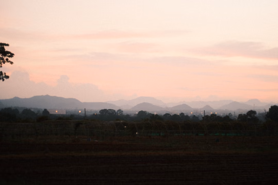Pink evening sky over a long mountain range, a dark field in the foreground with faint lights
