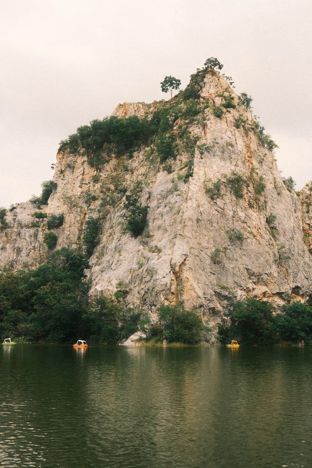 Single steep limestone crag rising above dark green lake water, tiny yellow pedal boats near its base