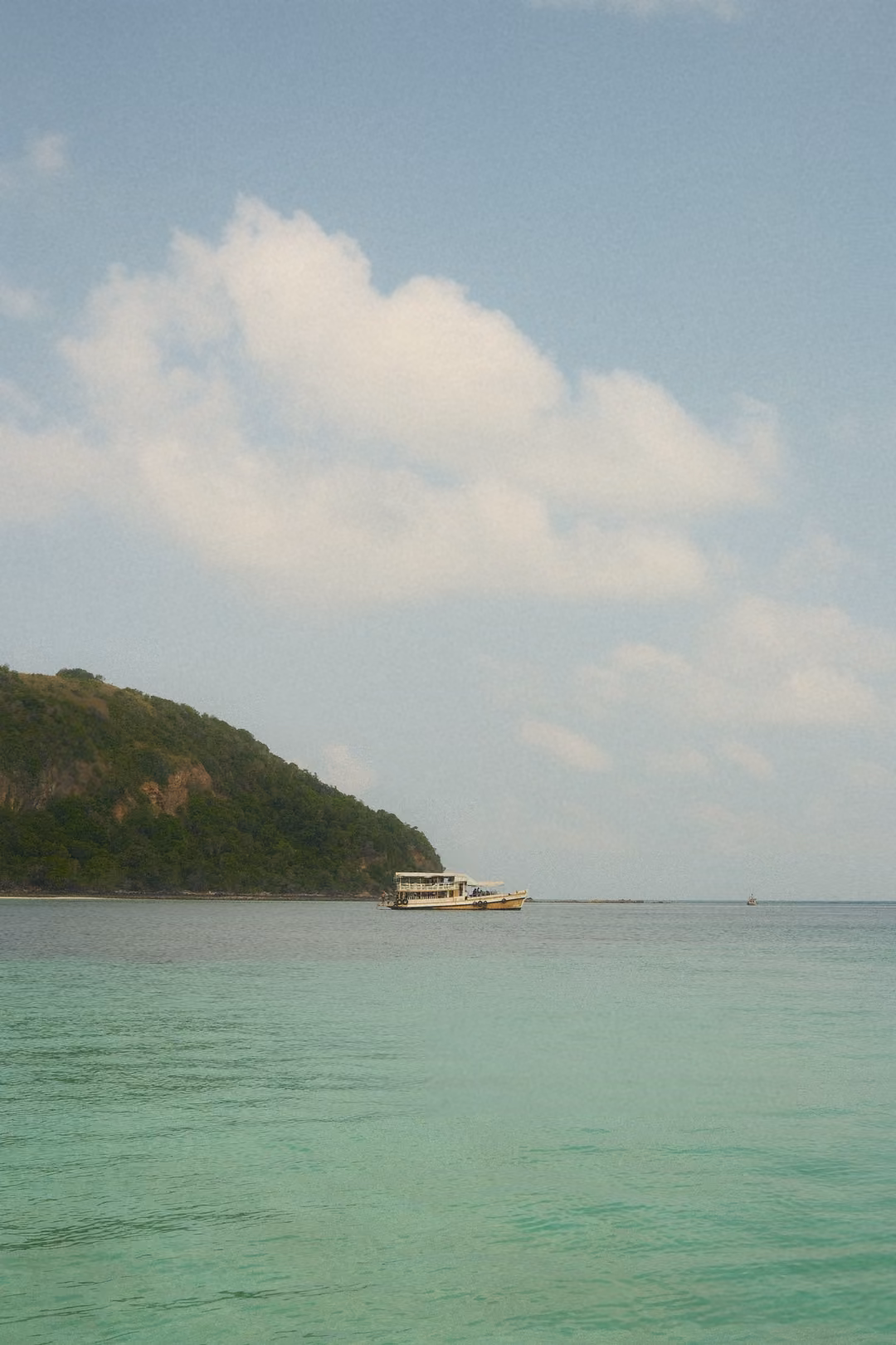 A wooded headland with a boat moored at its tip, turquoise sea and a bright blue sky with clouds.