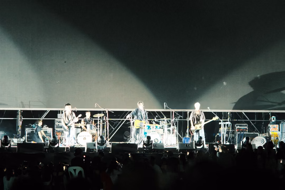 Band performing under two soft white light beams with a large lighting-rig shadow cast on the haze above