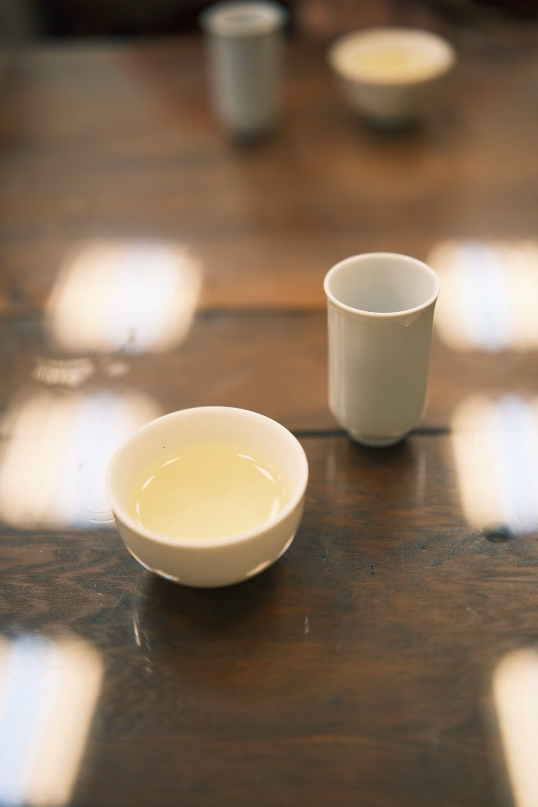 Bowl of light yellow tea and a white tasting cup on a polished dark table, another cup blurred behind