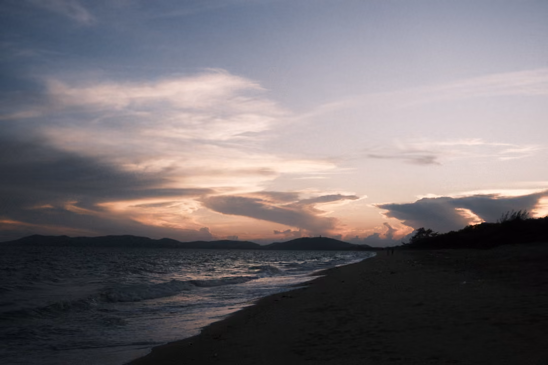 Dusky sky with lilac and pink clouds over a dark shoreline, waves and distant hills