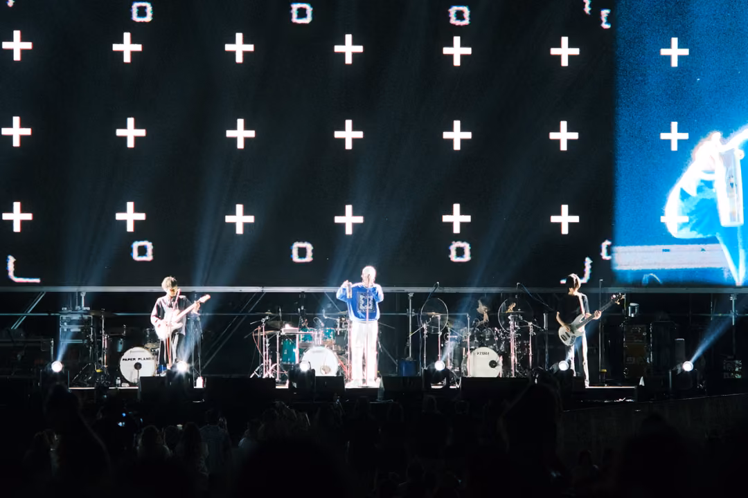 Band on stage below a screen grid of plus signs and squares, a blue close-up of the singer at the right edge.