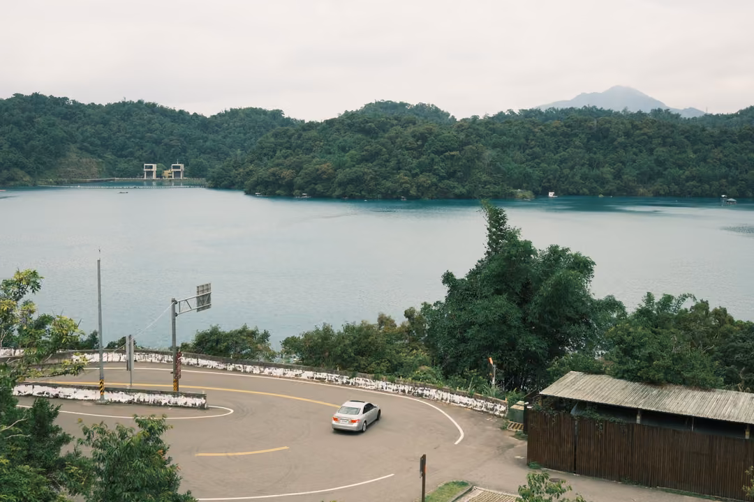 Single white car rounding a curved road beside a lake, wooded hills and a distant peak beyond