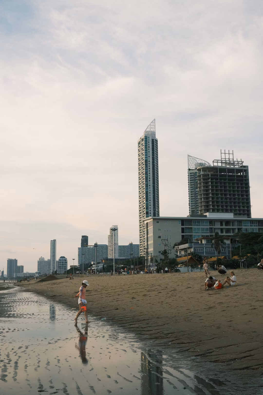 A small figure walking on wet sand near tall towers, people sitting on the beach beyond