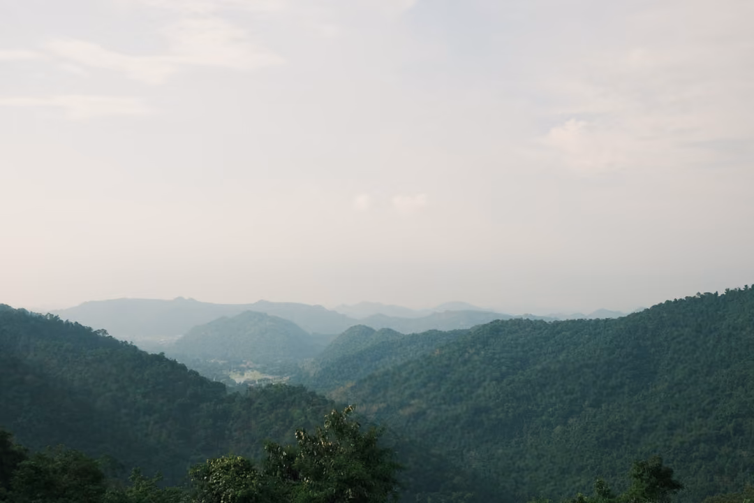 Layered forested mountain ridges fading into haze beneath a pale, cloudy sky.