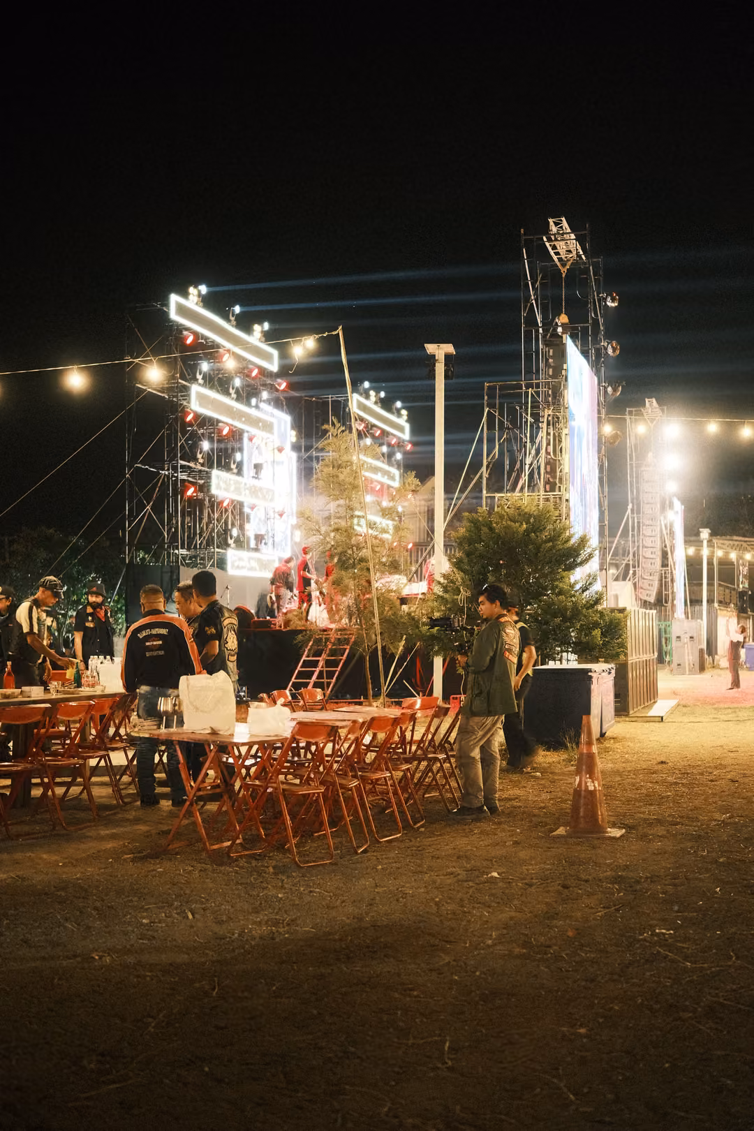 Night festival edge with a glowing light structure, red tables and chairs and a few people on dry grass
