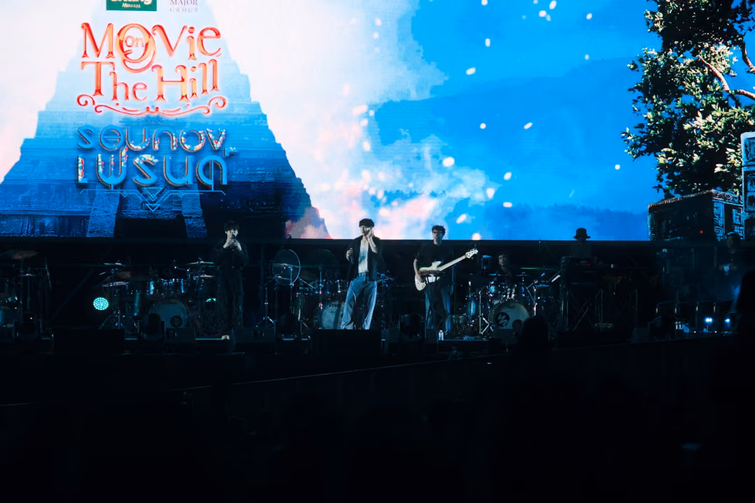 Stage with the Movie on The Hill logo on screen beside a temple-like silhouette and sparks, band in dim light below.